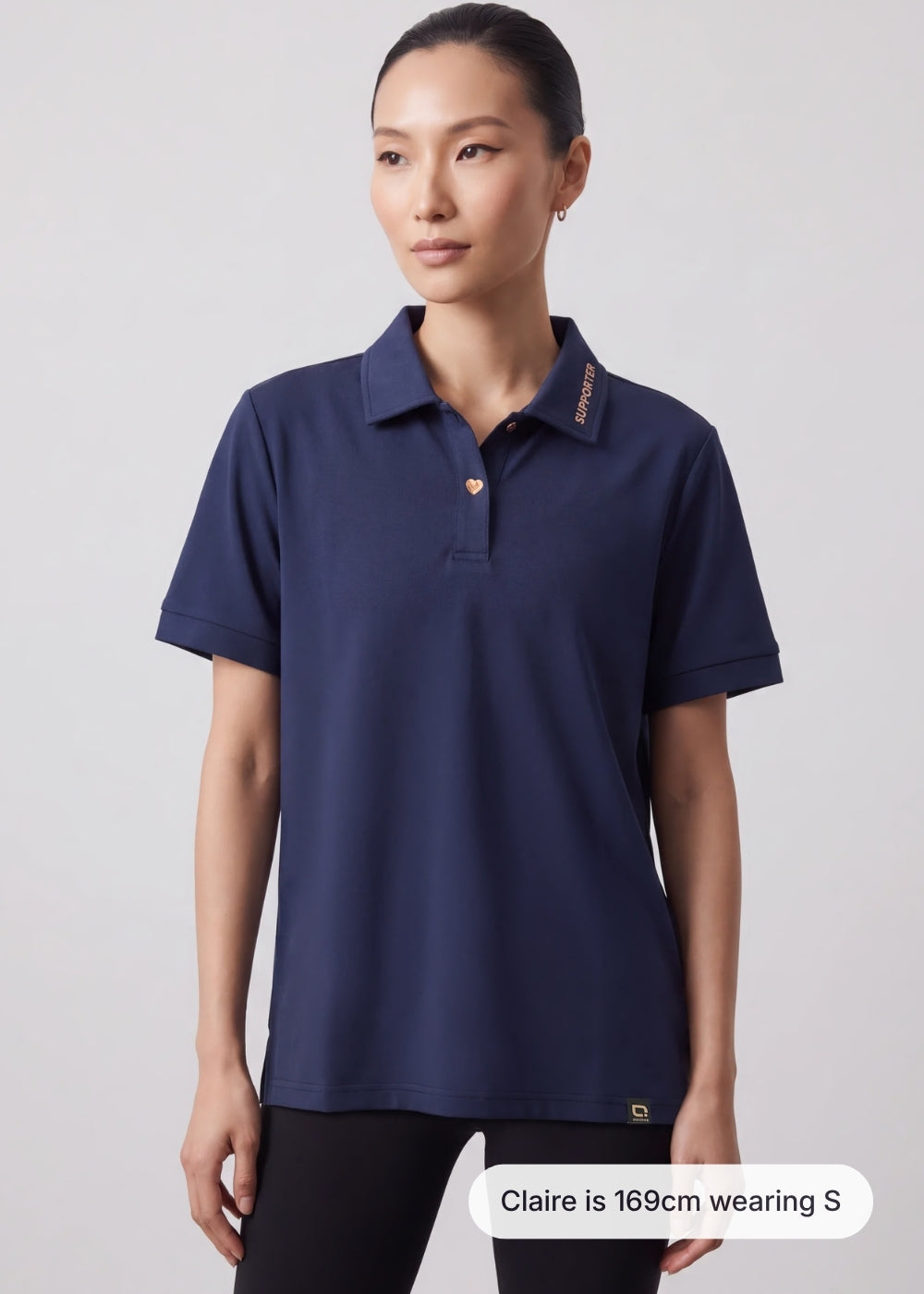 Model Claire wearing a navy blue women's polo shirt with 'SUPPORTER' collar text and heart buttons in size S