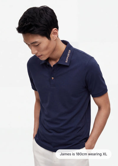 Model James wearing a navy blue men's polo shirt with 'SUPPORTER' collar text and white trousers in size XL.