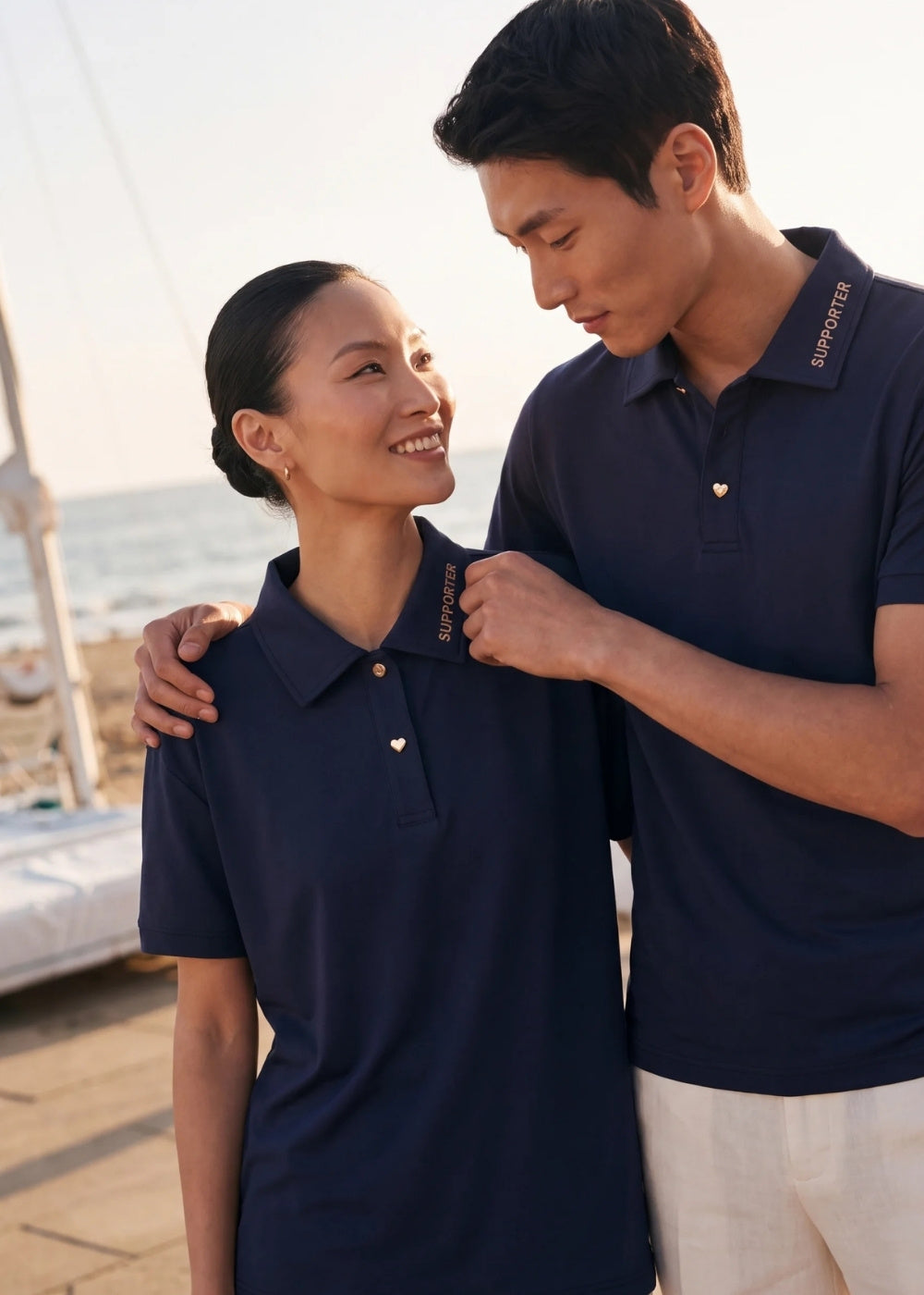 Couple wearing matching navy blue polo shirts with 'SUPPORTER' collar text and heart buttons by the beach.